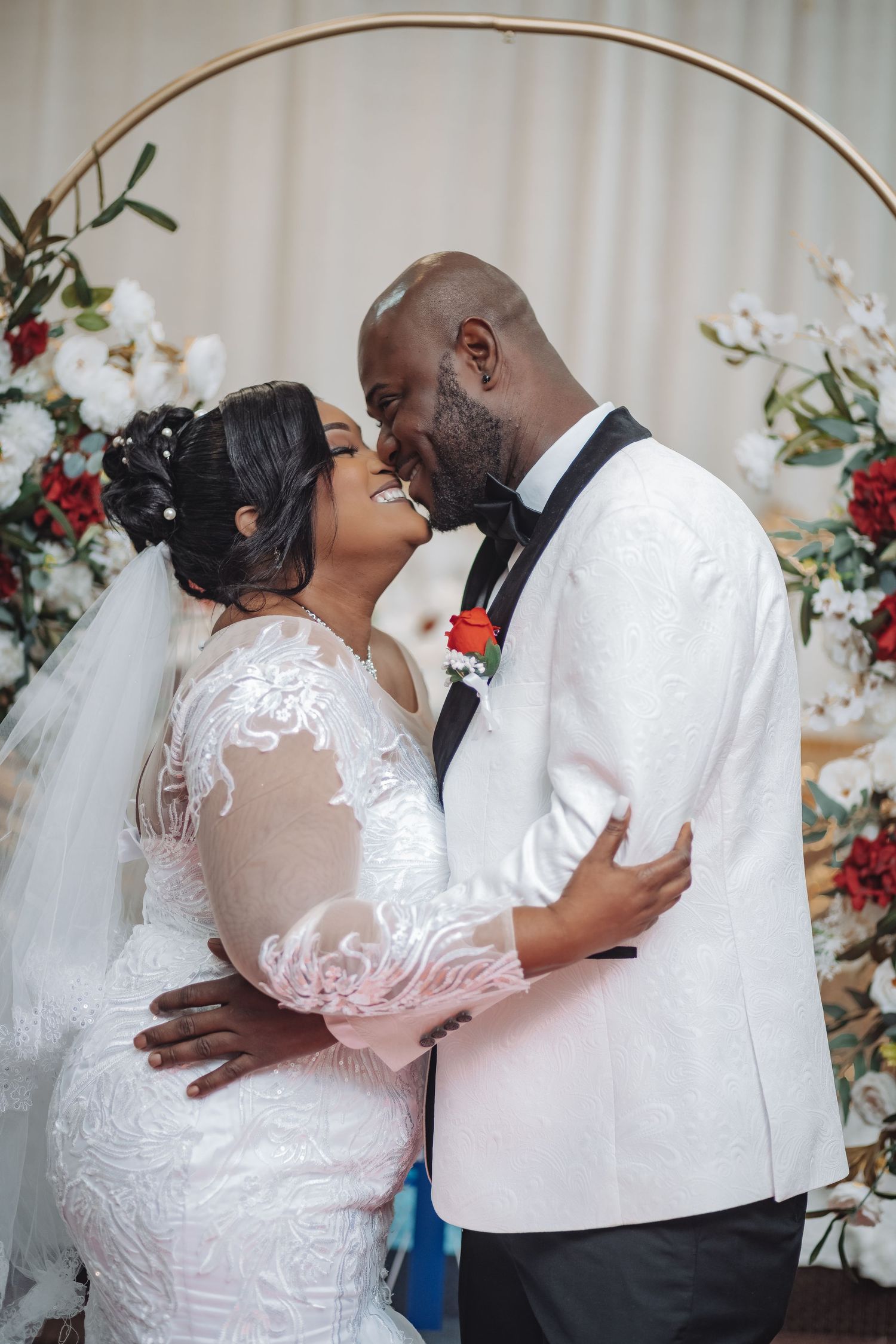A romantic wedding moment with a couple embracing under an arch decorated with red and white flowers.