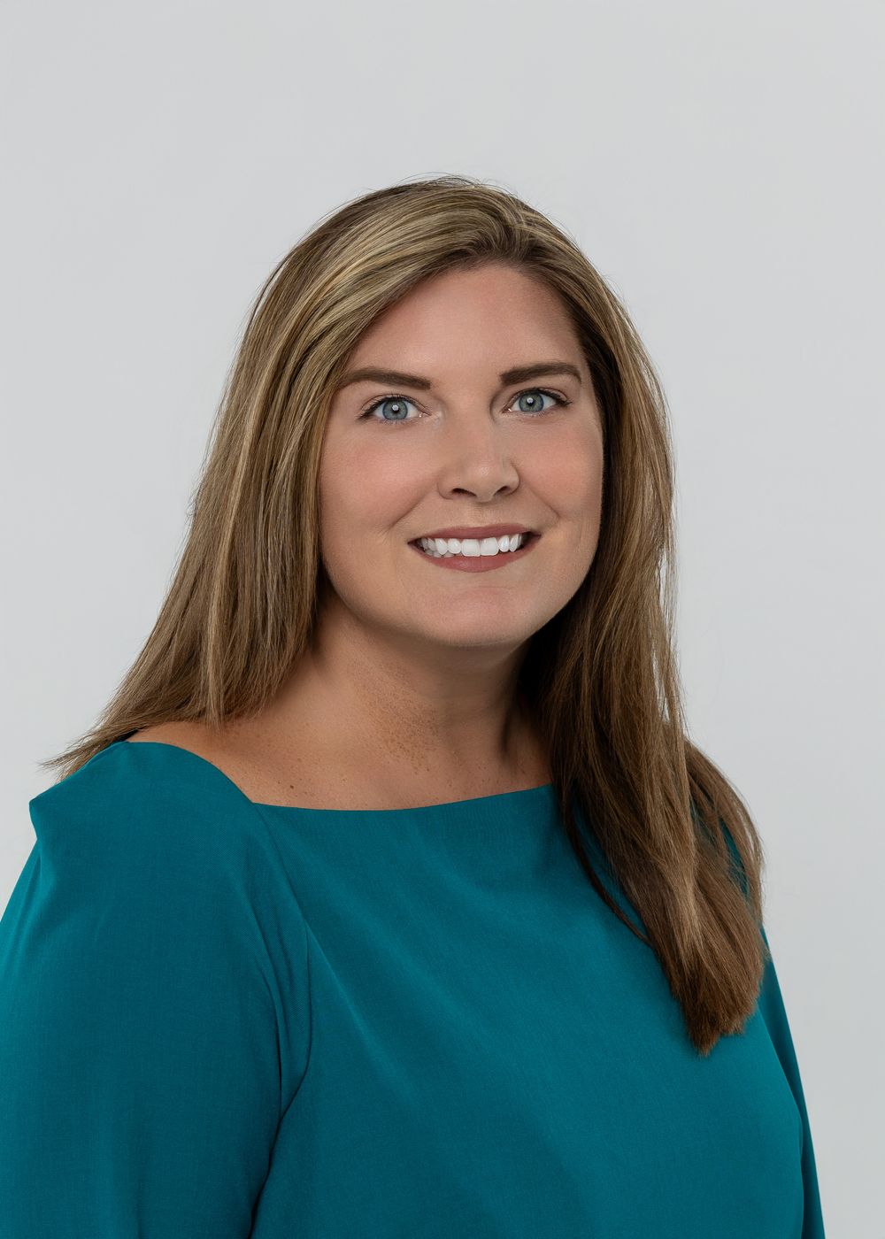 Professional headshot of a smiling woman with long blonde hair wearing a teal top against a gray background.