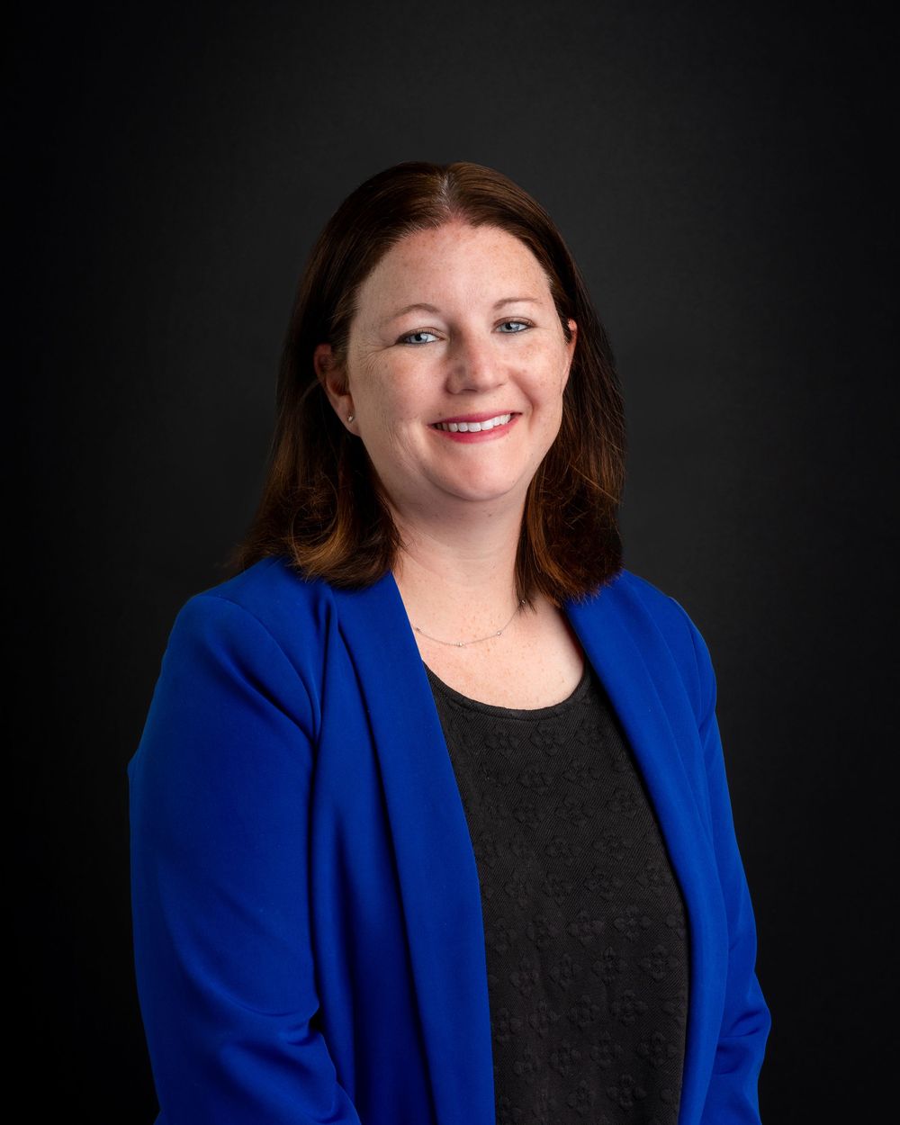 Professional headshot of a smiling woman with brown hair wearing a blue blazer against a dark background.