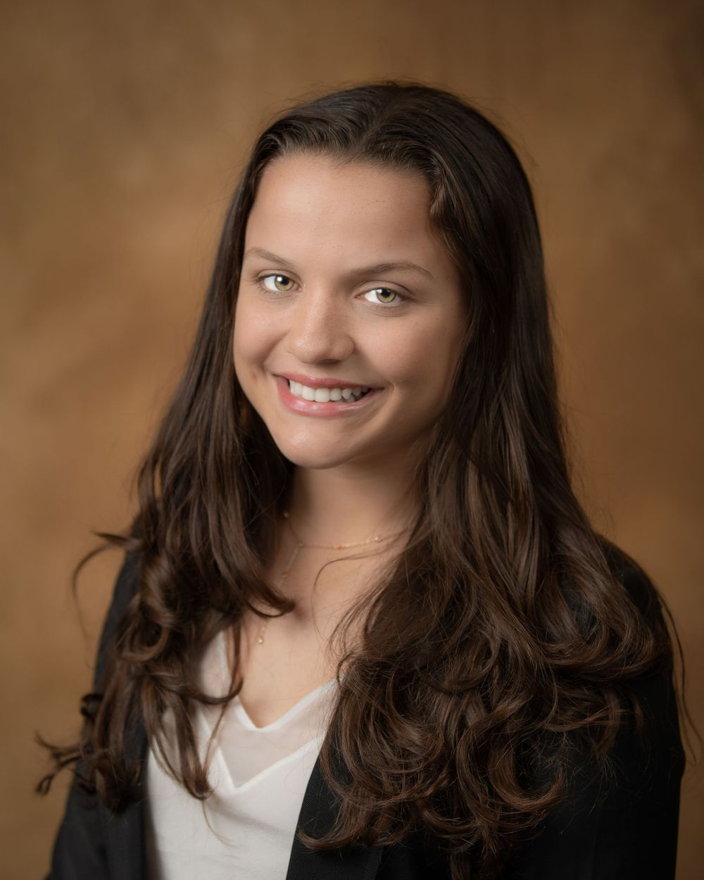Professional portrait of person with long dark hair wearing black blazer against brown background.