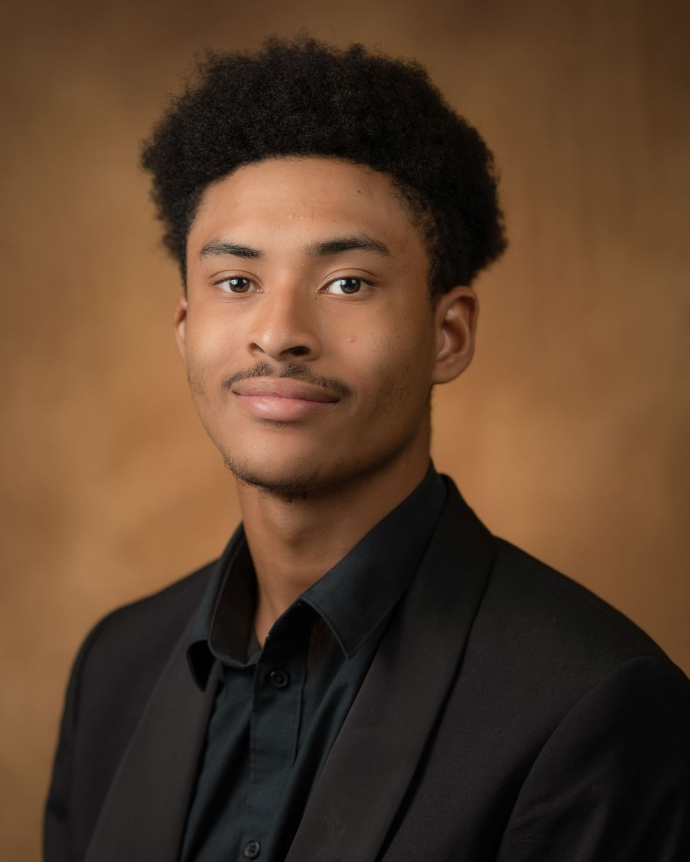 Professional headshot portrait against brown backdrop wearing black suit and shirt.