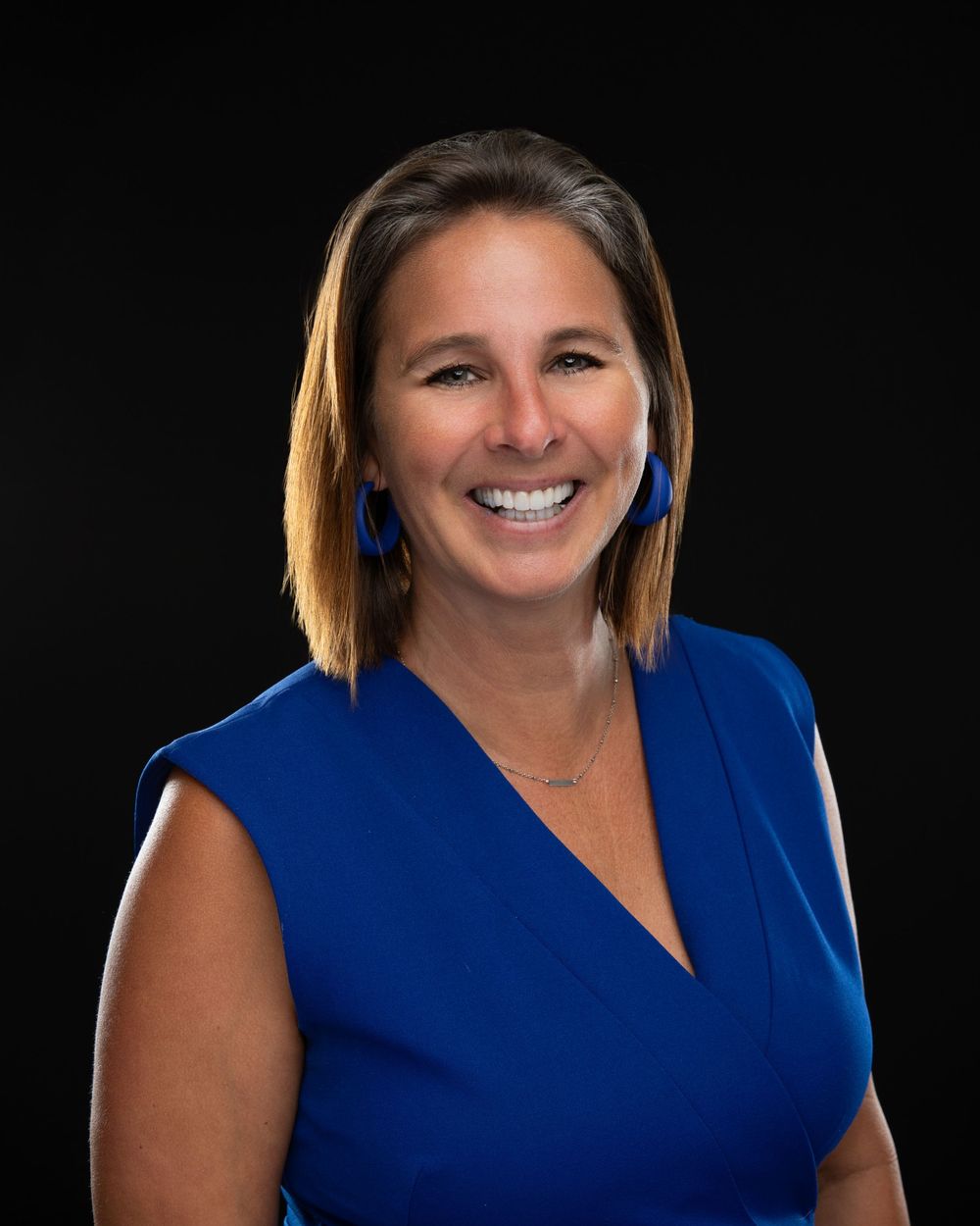 Professional headshot of a smiling woman in a blue sleeveless dress with blue earrings against a dark background.