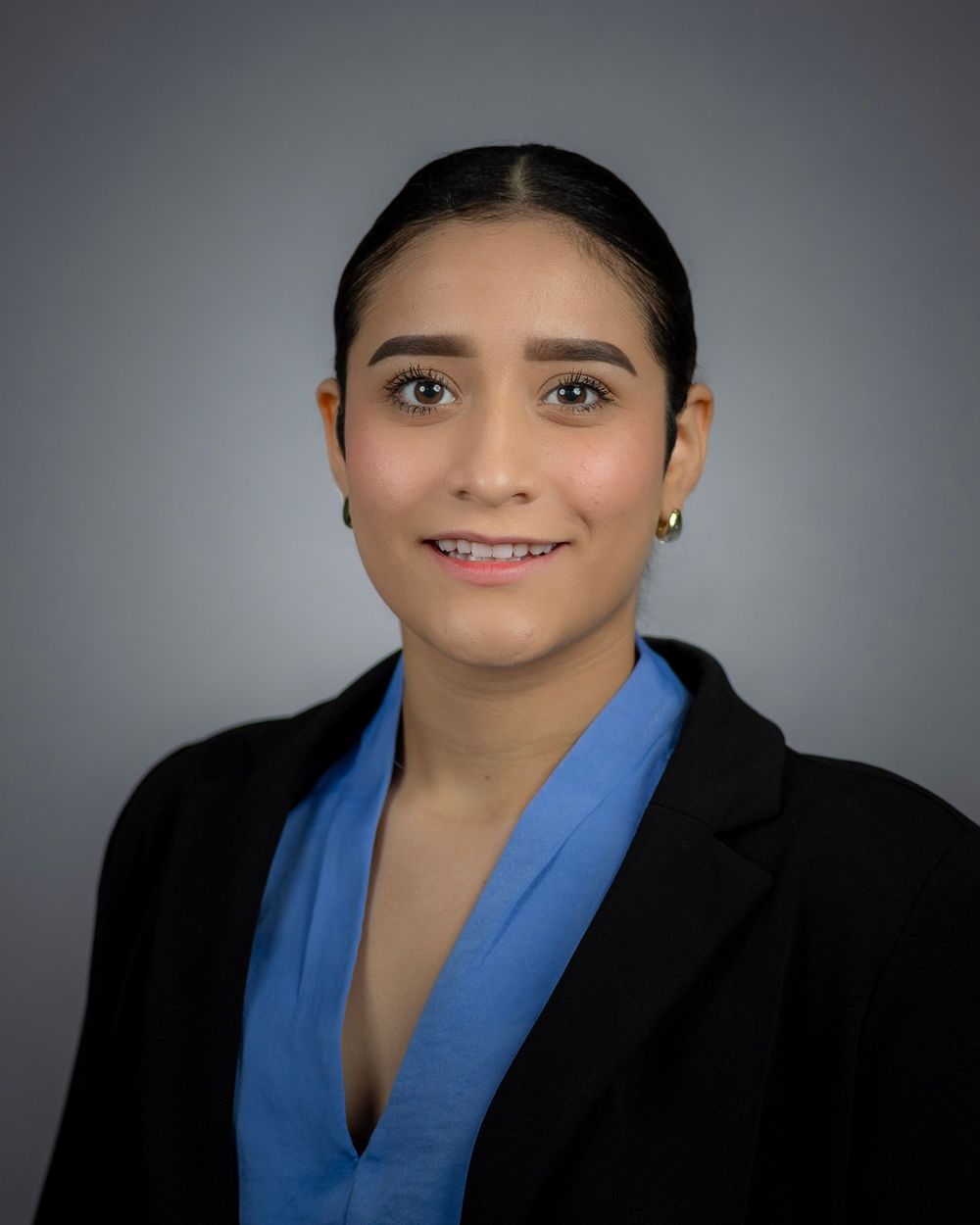 Professional headshot of person in black blazer and blue top against gray backdrop.