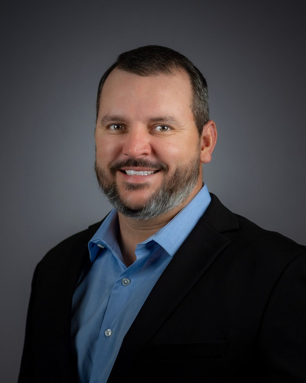 Professional headshot of person in black blazer and blue shirt against gray background.