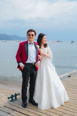 Wedding couple poses on wooden pier overlooking lake with mountains, bride in white and groom in burgundy jacket.