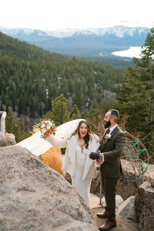 Wedding couple laughs together while standing on rocky mountain overlook with snowy peaks in background.
