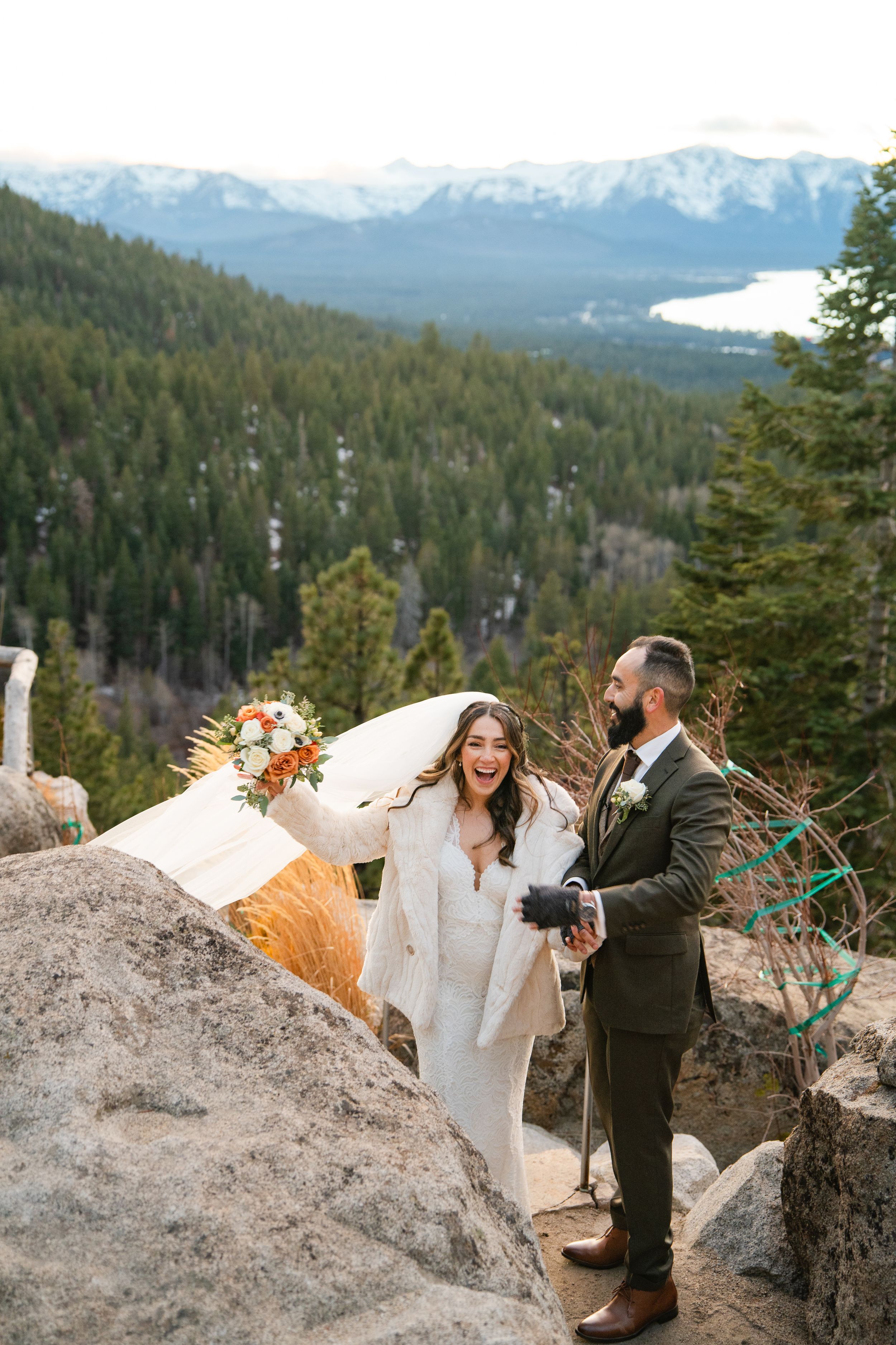 Wedding couple laughs together while standing on rocky mountain overlook with snowy peaks in background.