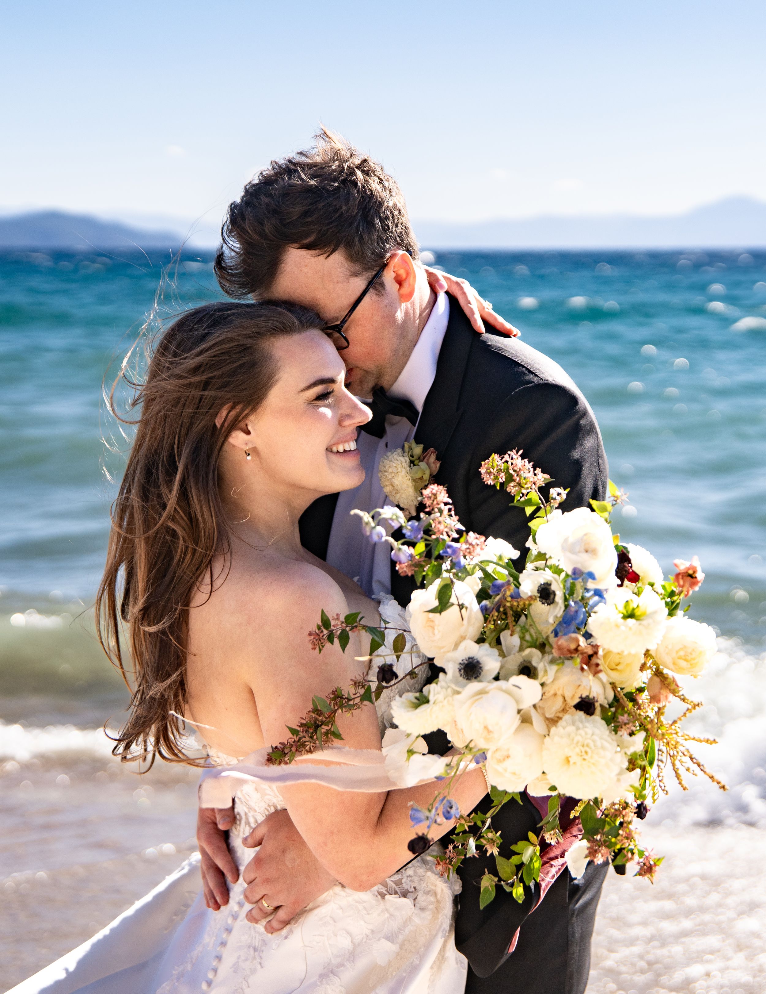 A couple embraces on a beach with turquoise waters, holding a beautiful bouquet of white and blue flowers.