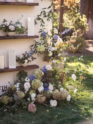 Rustic floral arrangements with white hydrangeas and blue blooms cascading along wooden shelves against a cream wall.