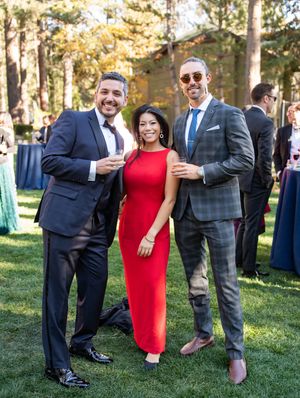 Three guests in formalwear pose outdoors at an outdoor wedding reception with trees in the background.