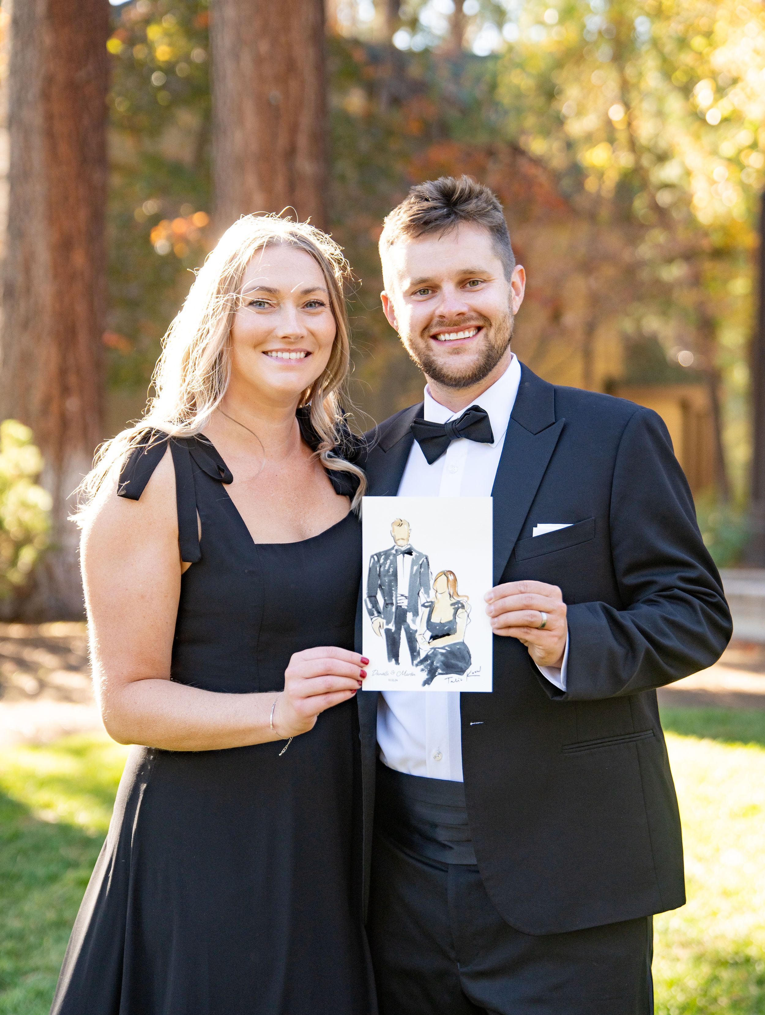 A couple in formal attire stands outdoors holding a black and white photo in a sunlit wooded setting.