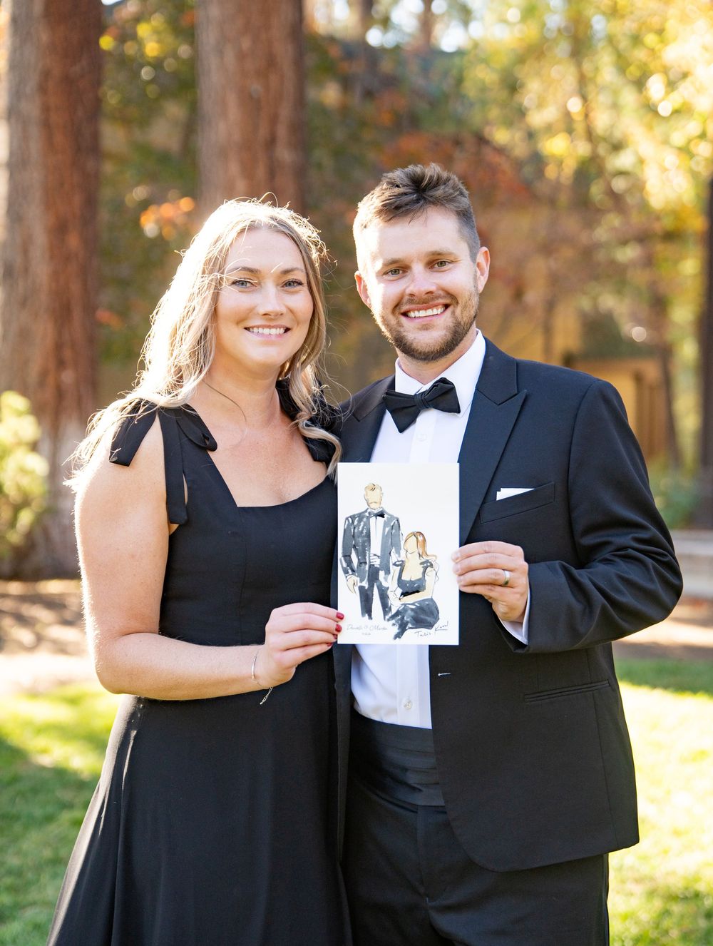 A couple in formal attire stands outdoors holding a black and white photo in a sunlit wooded setting.