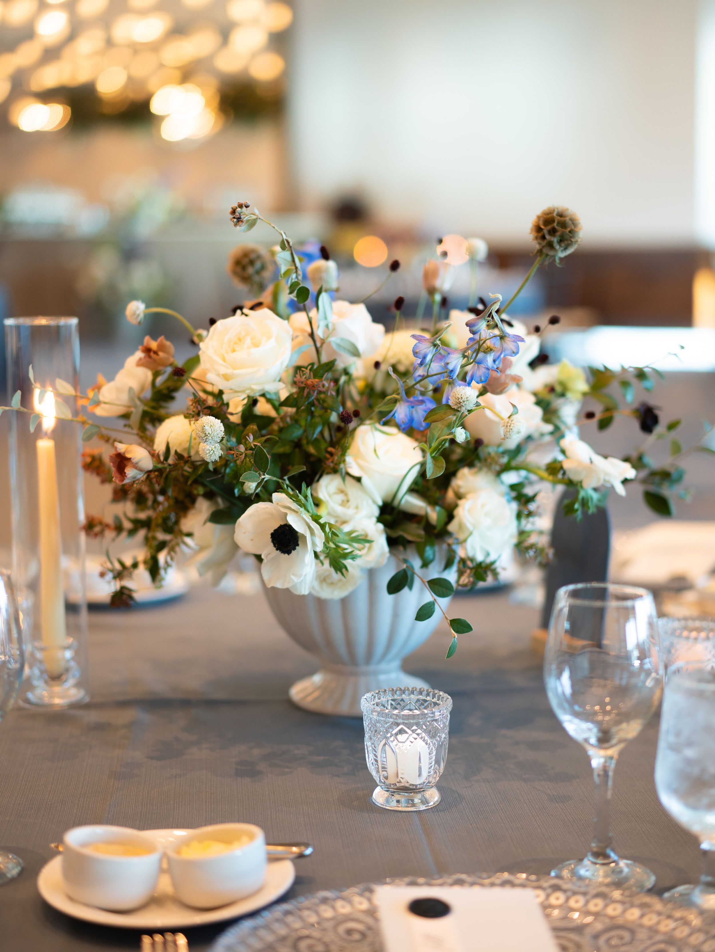 Elegant white floral centerpieces with roses and delphinium arranged in ceramic vases line a neutral tablescape.