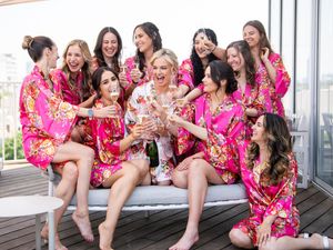 Group wearing matching pink floral robes gather for celebratory pre-wedding moment on balcony.