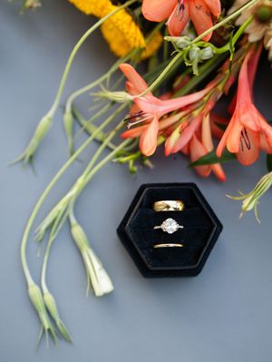 Engagement and wedding rings displayed in black hexagonal box with coral flowers and greenery on gray background.