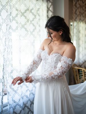 Bride wearing an elegant off-shoulder white lace wedding gown with sheer long sleeves stands near a window with patterned curtains.