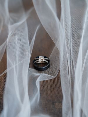 Wedding rings displayed on delicate white tulle fabric with a princess cut diamond engagement ring and black band.