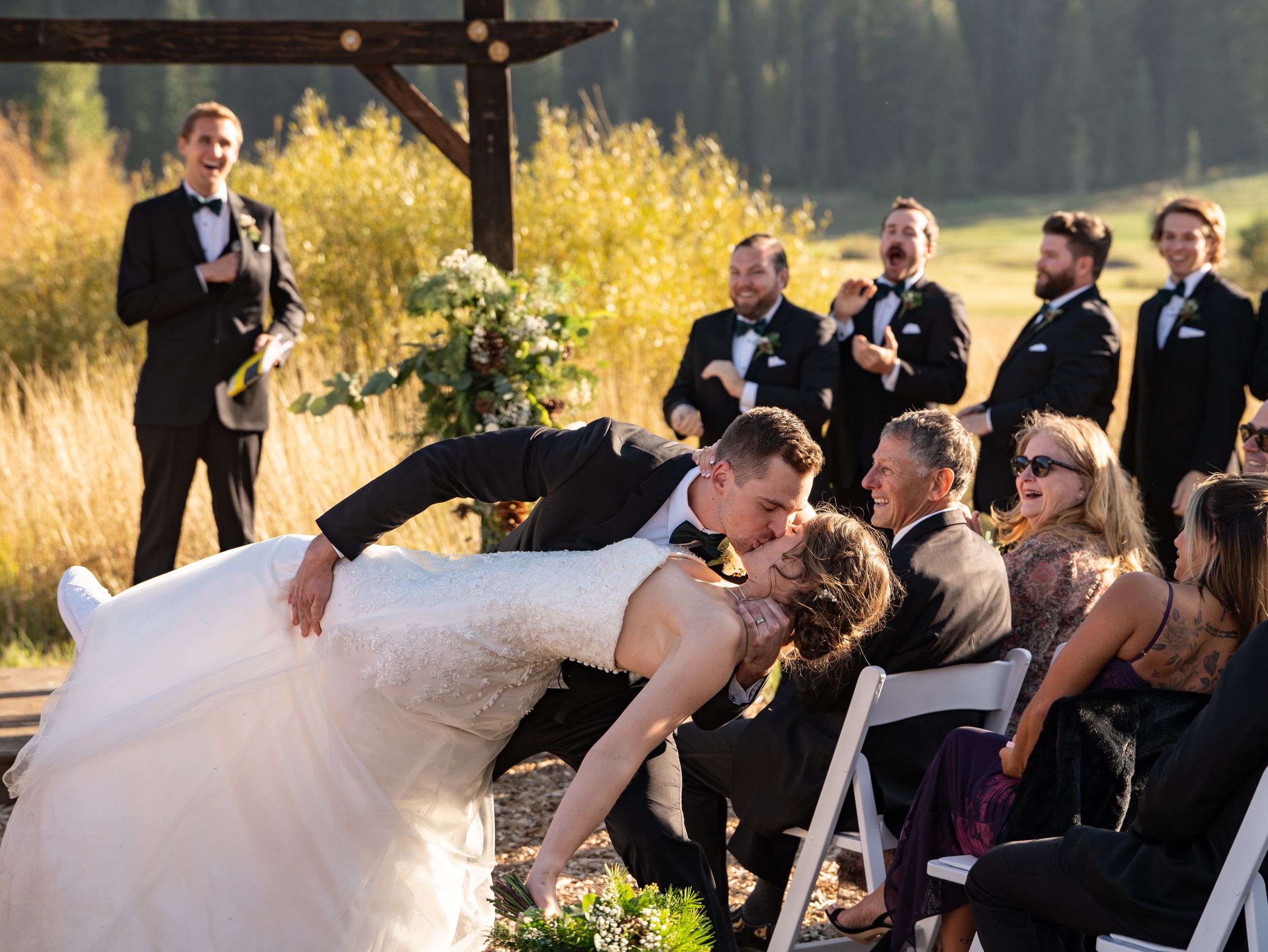 A wedding ceremony at sunset with a dramatic dip kiss between newlyweds while guests and wedding party look on.