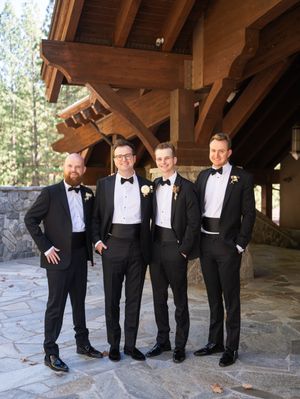 Groomsmen wearing classic black tuxedos pose together under a rustic stone pavilion in a playful series of group shots.