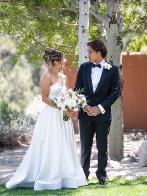 Newlywed couple in formalwear stands together outdoors with birch trees, bride holding white and blush floral bouquet.