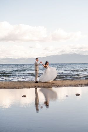 Couple dances on beach at sunset with reflection in water and mountains in background.
