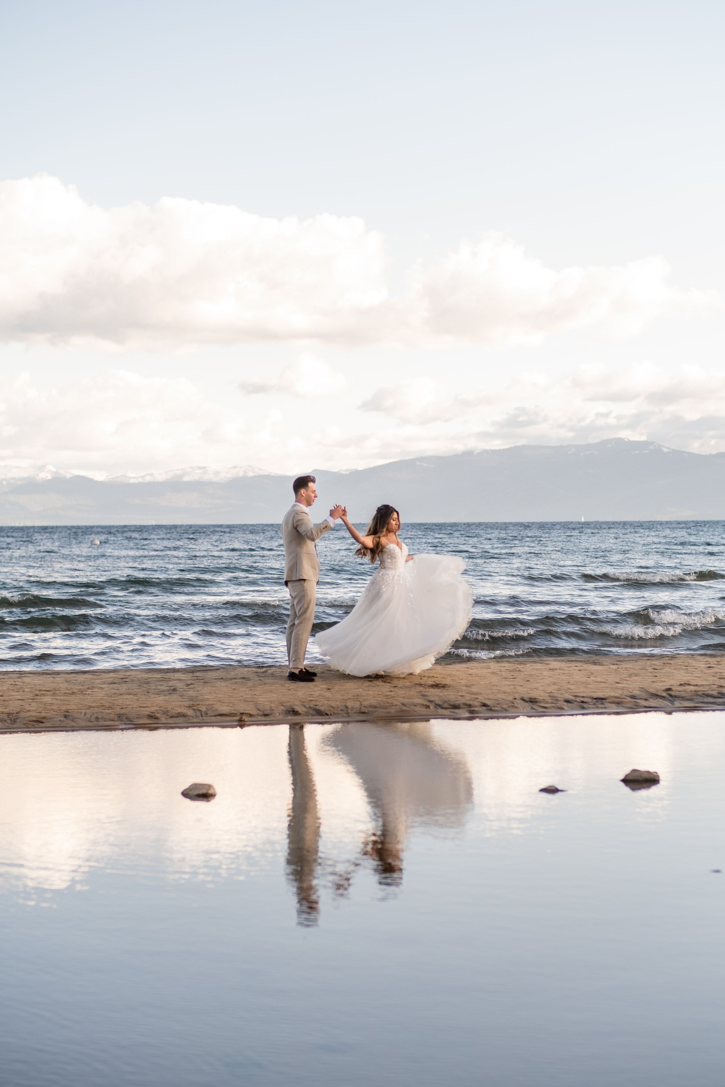 Couple dances on beach at sunset with reflection in water and mountains in background.