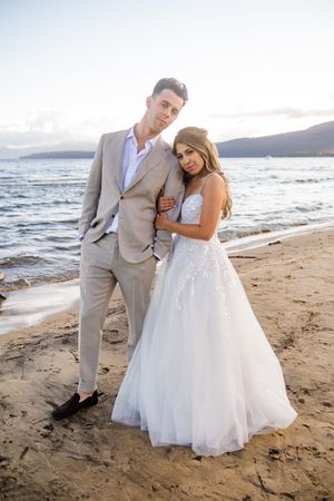 A couple shares romantic moments on a beautiful beach at sunset in wedding attire, with mountains in the background.