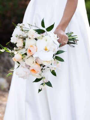 Close-up of elegant white and peach floral bouquets with greenery held against flowing white wedding dress fabric.