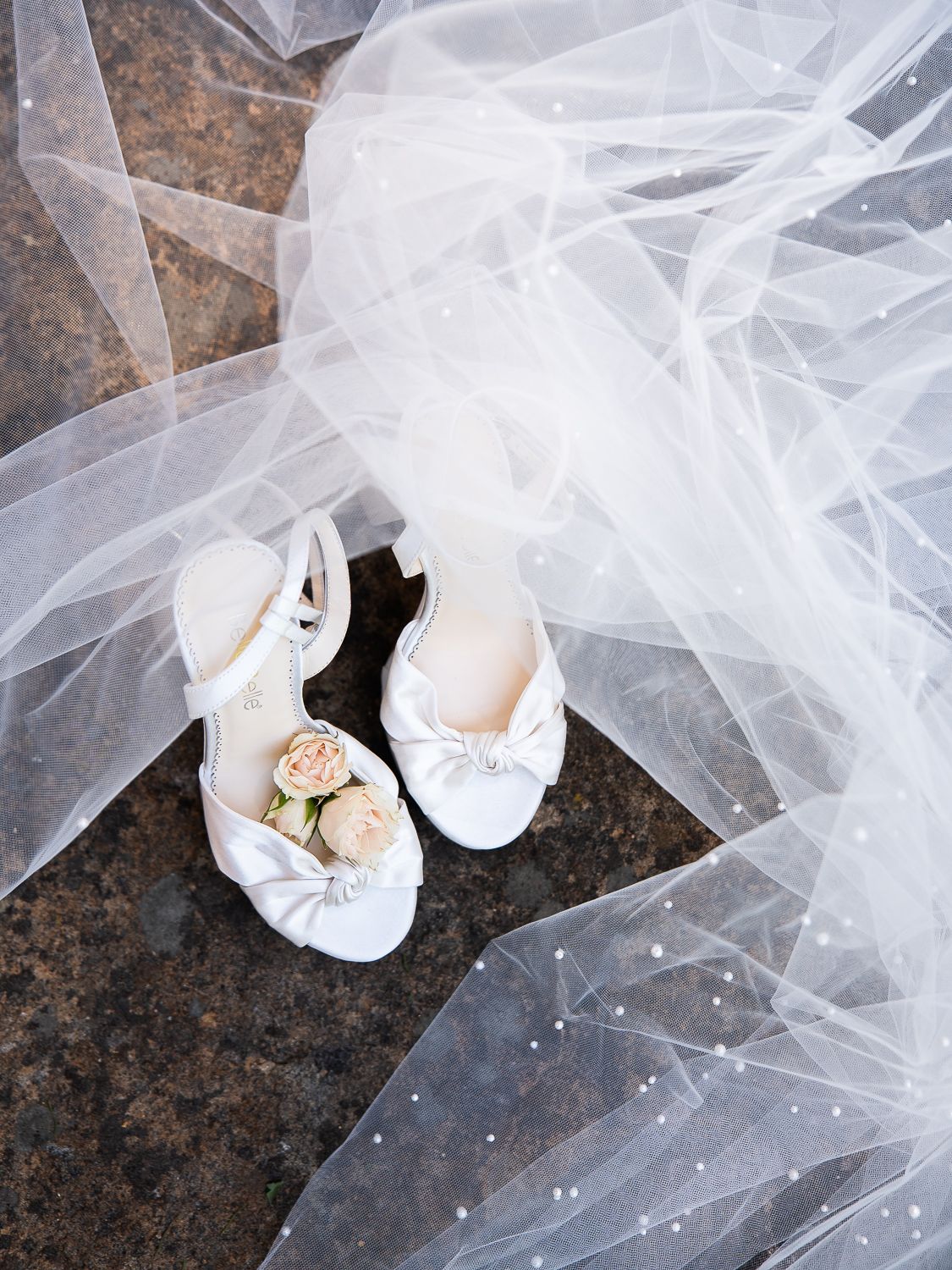 White bridal heels with bow detail and blush roses rest on marble floor beside a flowing pearl-trimmed wedding veil.