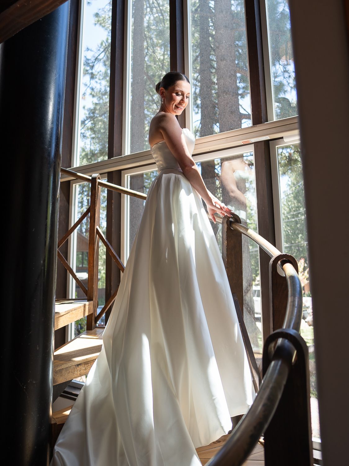 Bride in elegant white ball gown smiling on staircase with large windows and forest views behind her.