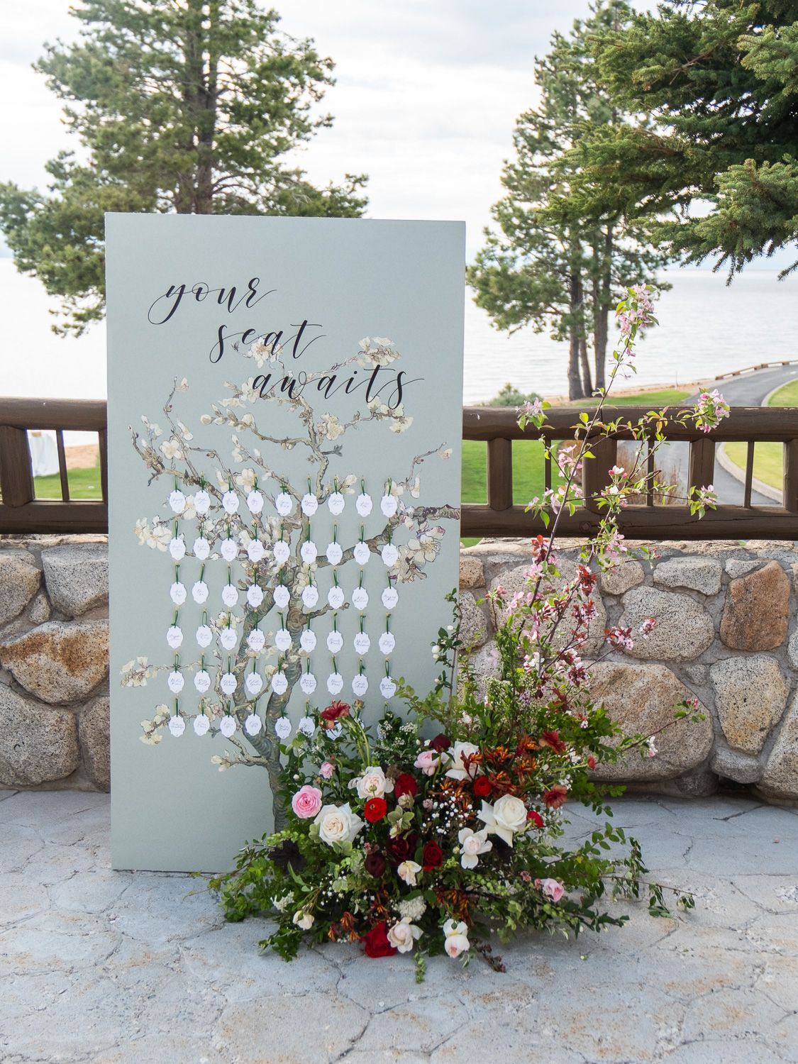Elegant wedding seating chart display board with floral arrangement, reading your seat awaits in calligraphy script outdoors.