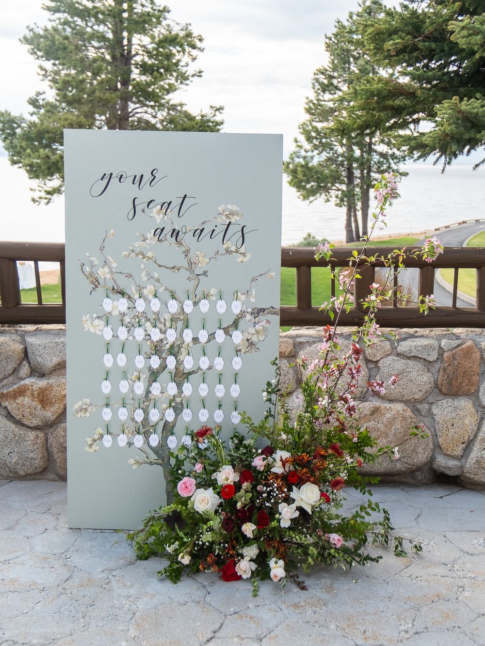 Elegant wedding seating chart display board with floral arrangement, reading your seat awaits in calligraphy script outdoors.