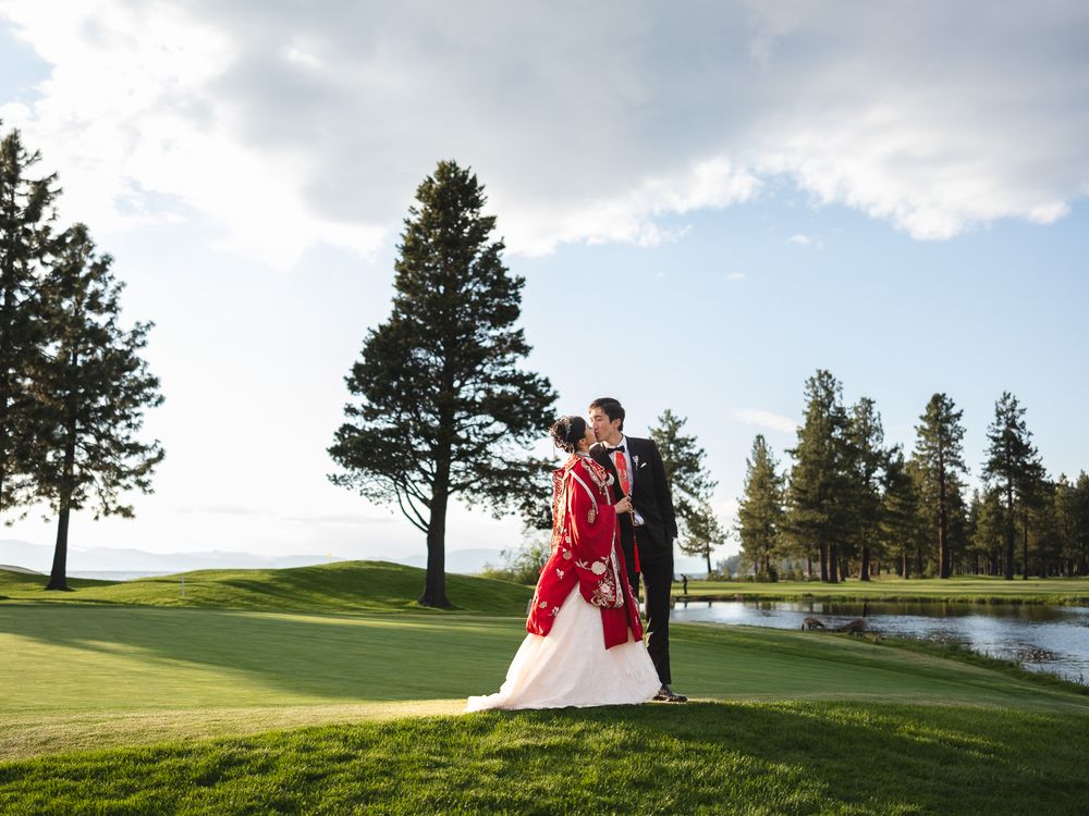 Bride in red gown and groom embrace on lush golf course with tall pines and blue sky backdrop.