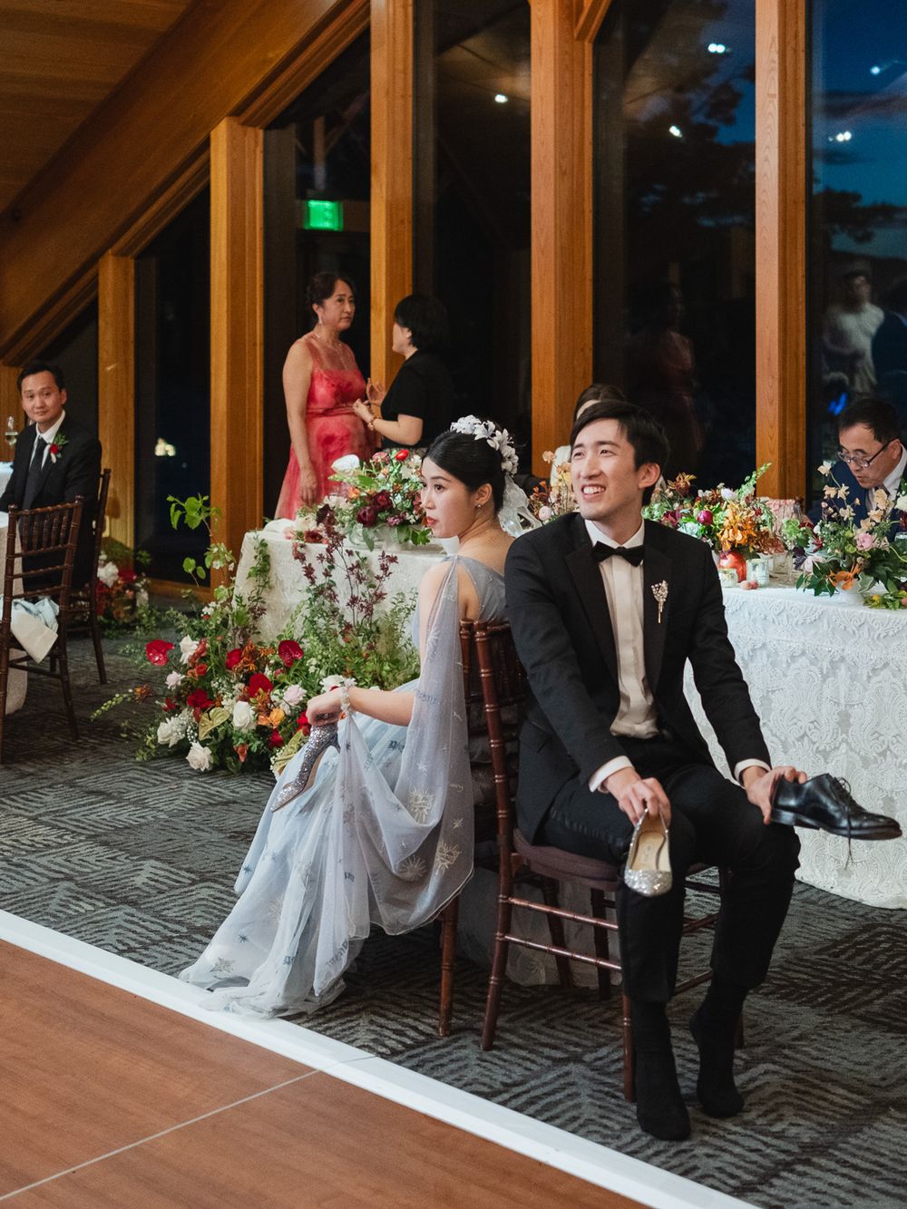 Asian bride and groom sit at sweetheart table adorned with red floral arrangements at elegant reception.