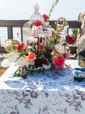 Elegant floral centerpiece with white birdcage, red and pink roses, pearls on floral tablecloth near waterfront.