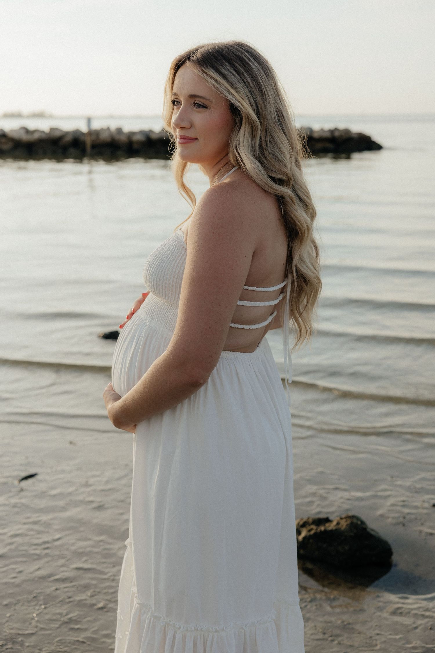 Woman with long dark hair poses outdoors in denim and white top against a coastal landscape at sunset.