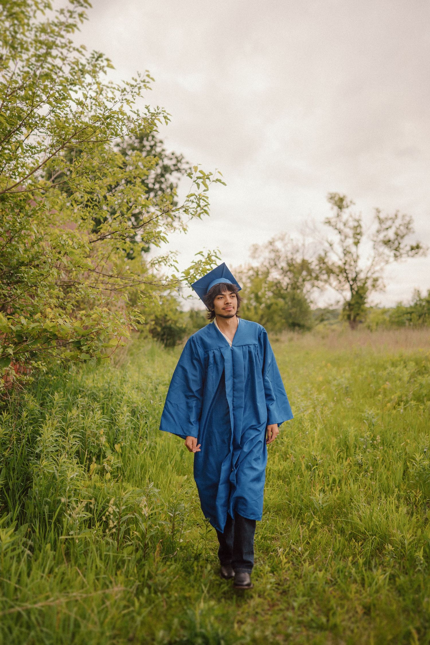 Small figure in bright blue coat walks through tall green grass in a meadow under cloudy skies.