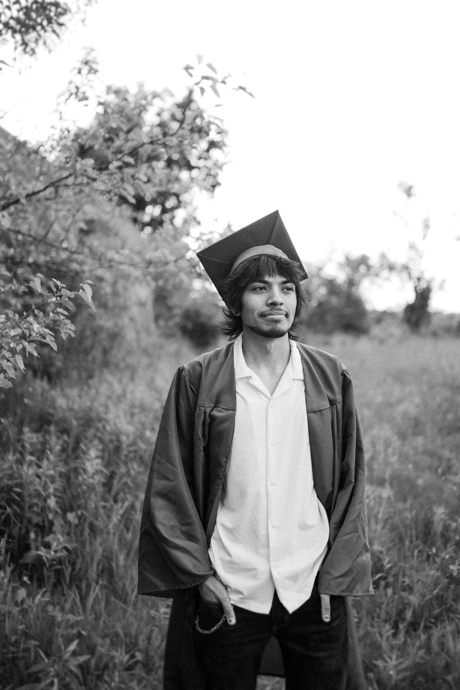 Graduate stands in a natural outdoor setting wearing academic regalia and white shirt in black and white photograph.