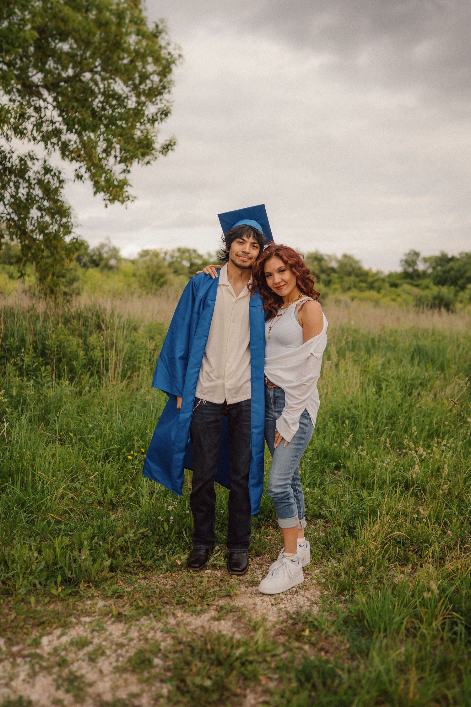 A couple stands together in a grassy field on an overcast day, with one wearing blue graduation attire.