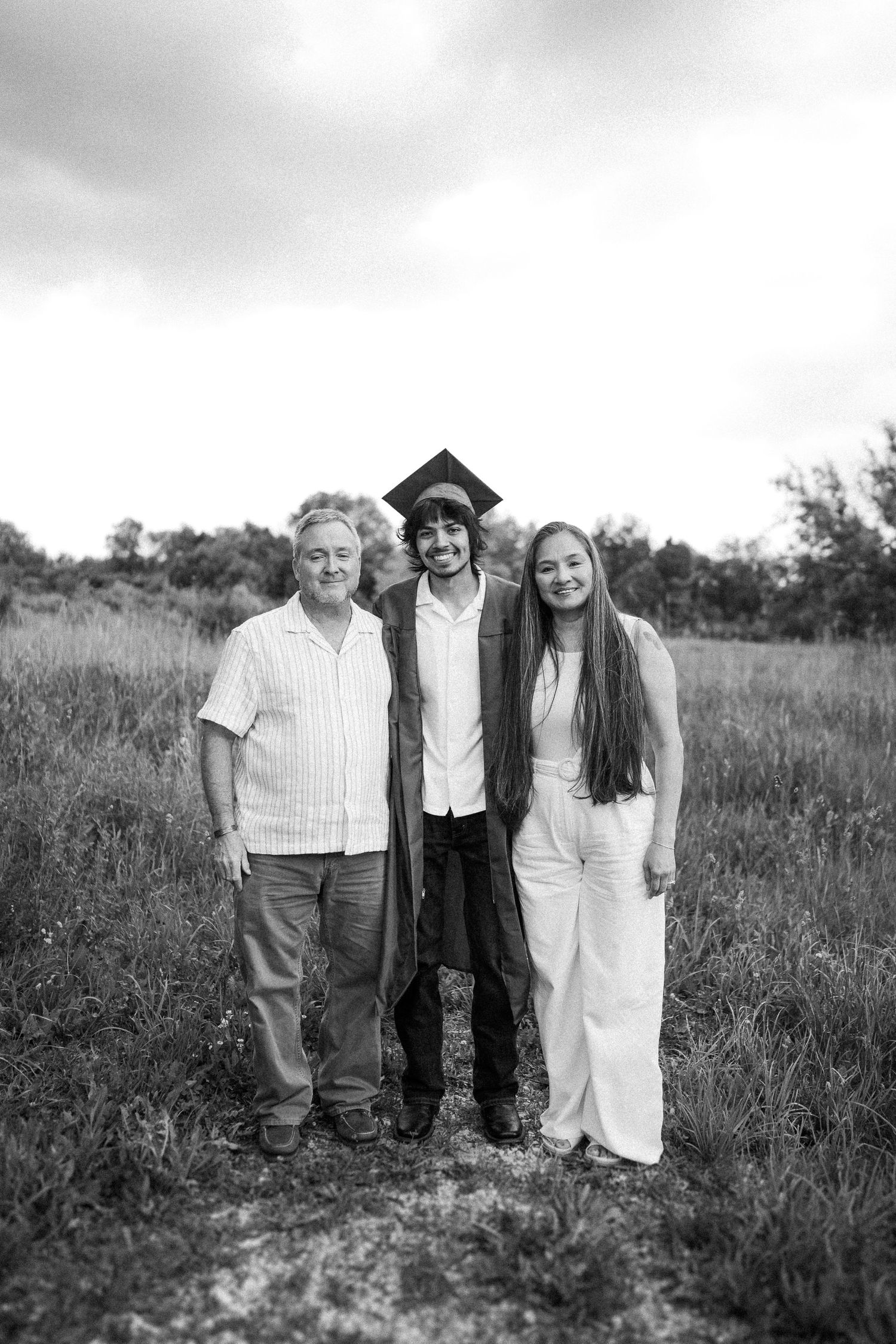 Black and white graduation photo showing three people standing together in a grassy field outdoors.
