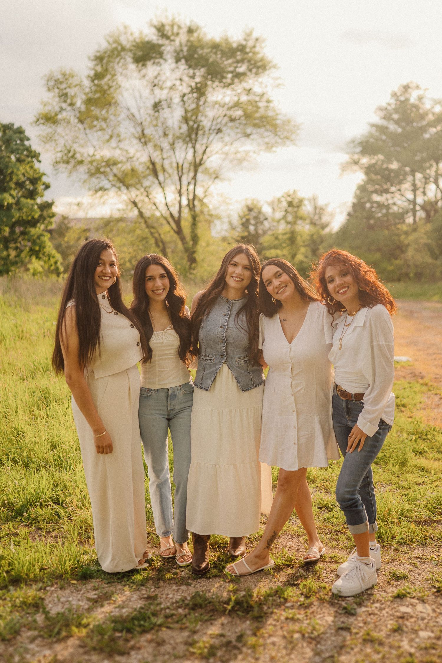 Five friends in coordinated white and denim outfits pose together in a sunlit field at golden hour.