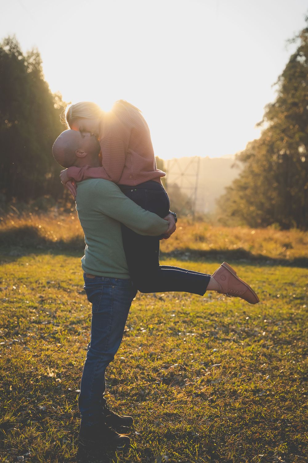 sunset glow in the background and light shines through a couples kiss while the man lifts the woman in a loving embrace