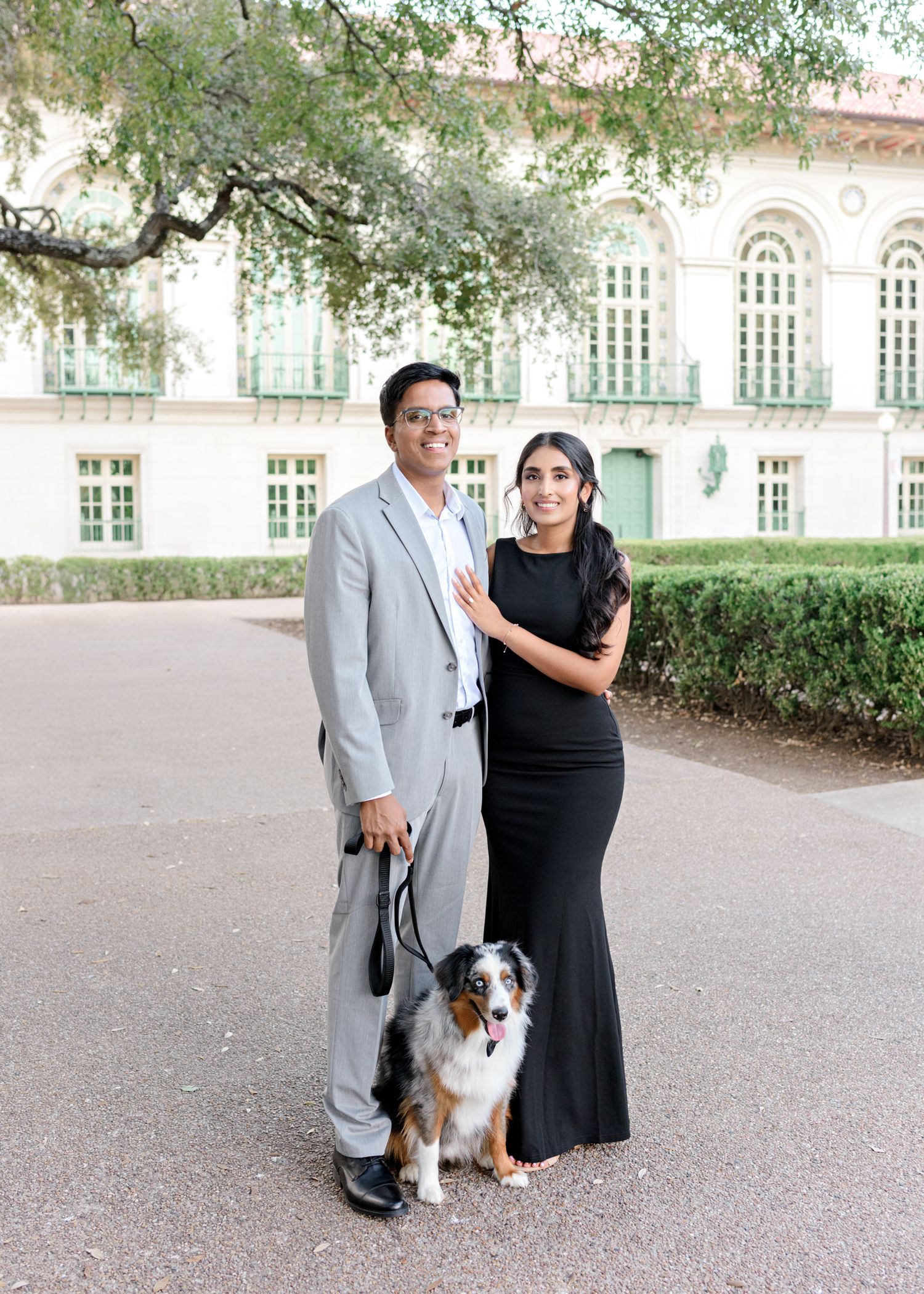 A couple poses with their Australian Shepherd dog in front of an elegant building with arched windows and greenery.