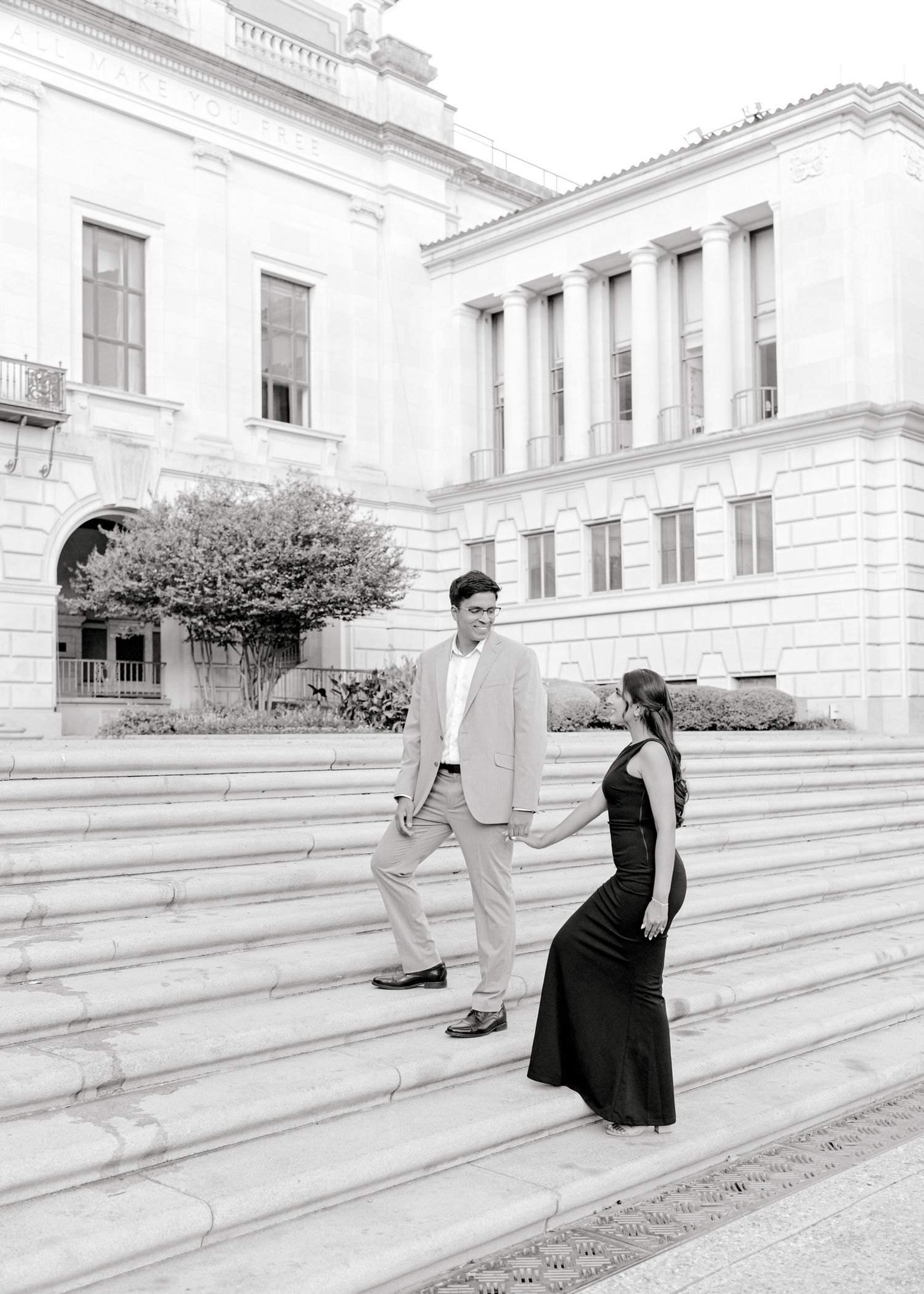Couple in formal attire playfully interacts on grand white stone steps of historic building in black and white series.