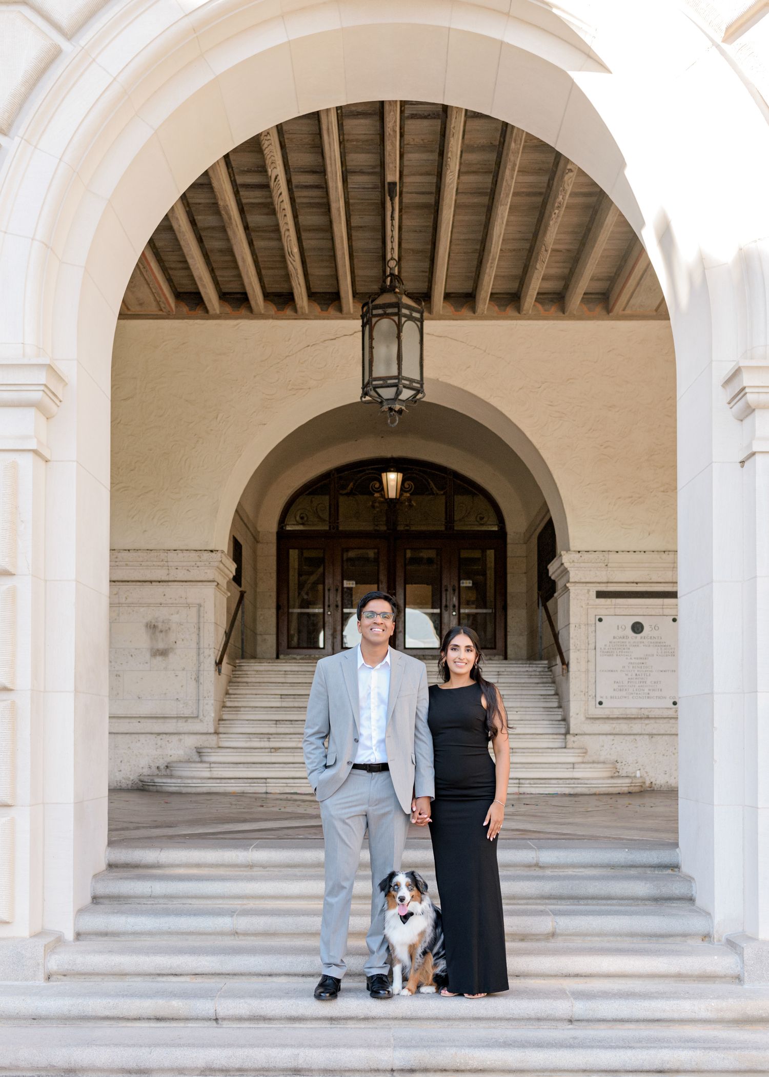 A couple and their Australian Shepherd dog pose on grand white stone steps beneath an elegant arched entrance.