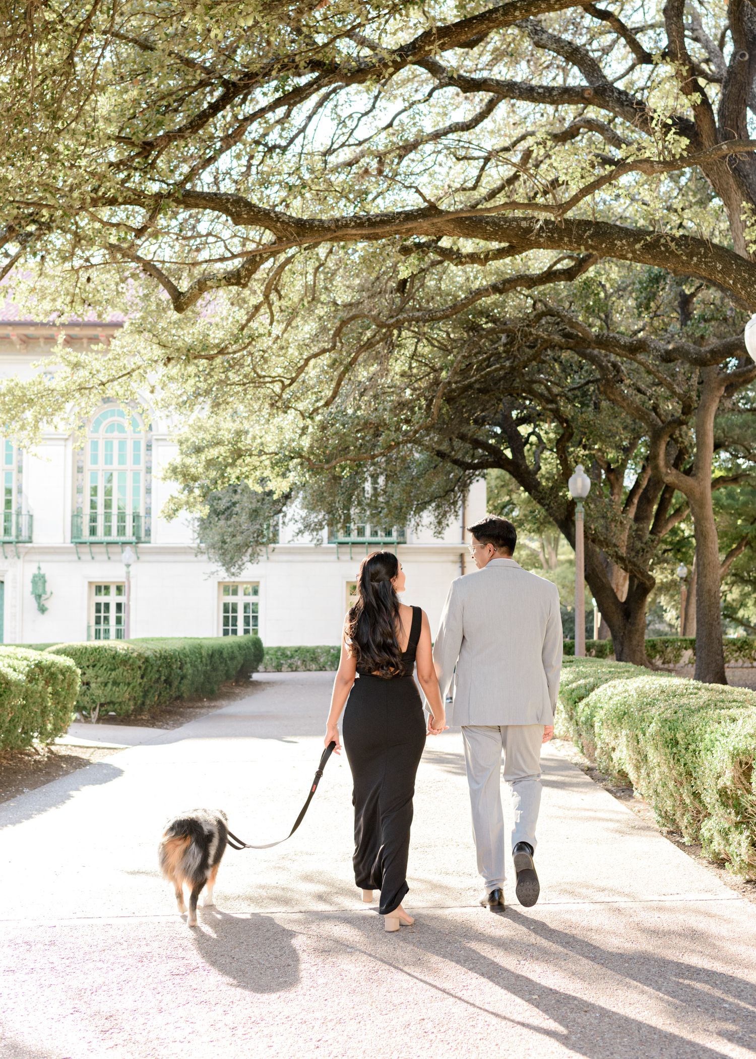 Couple walking with their dog under moss-draped oak trees on a sunny path by a grand historic building.