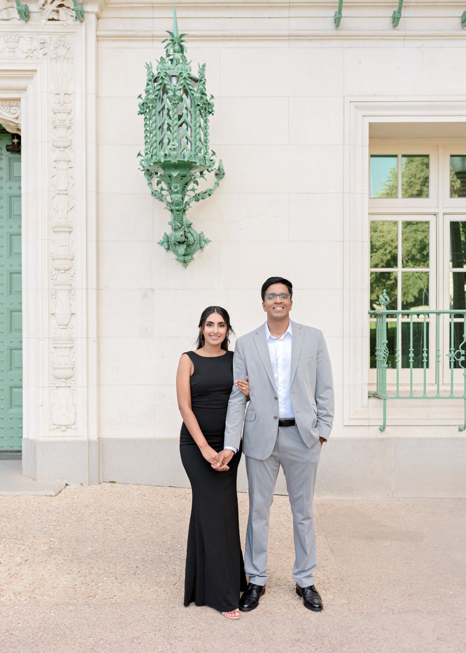 A couple poses in front of a white building with elegant green architectural details and shutters.