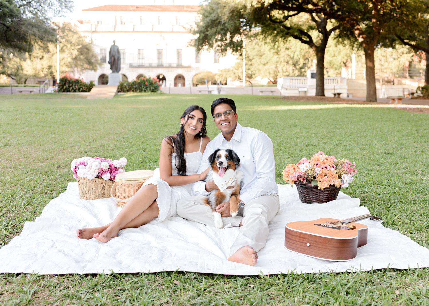 A couple enjoys a romantic picnic with flowers and snacks on a blanket in a grassy field.