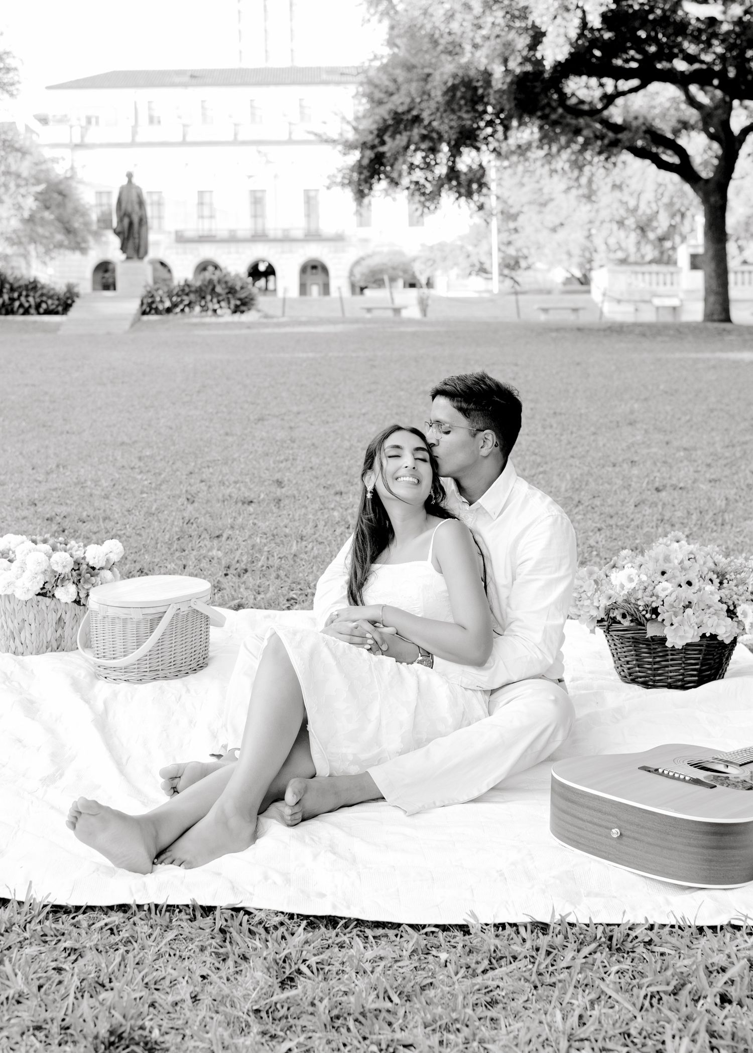 Romantic outdoor picnic setup on grass with white blanket, guitar case, and flower baskets in a black and white photo.