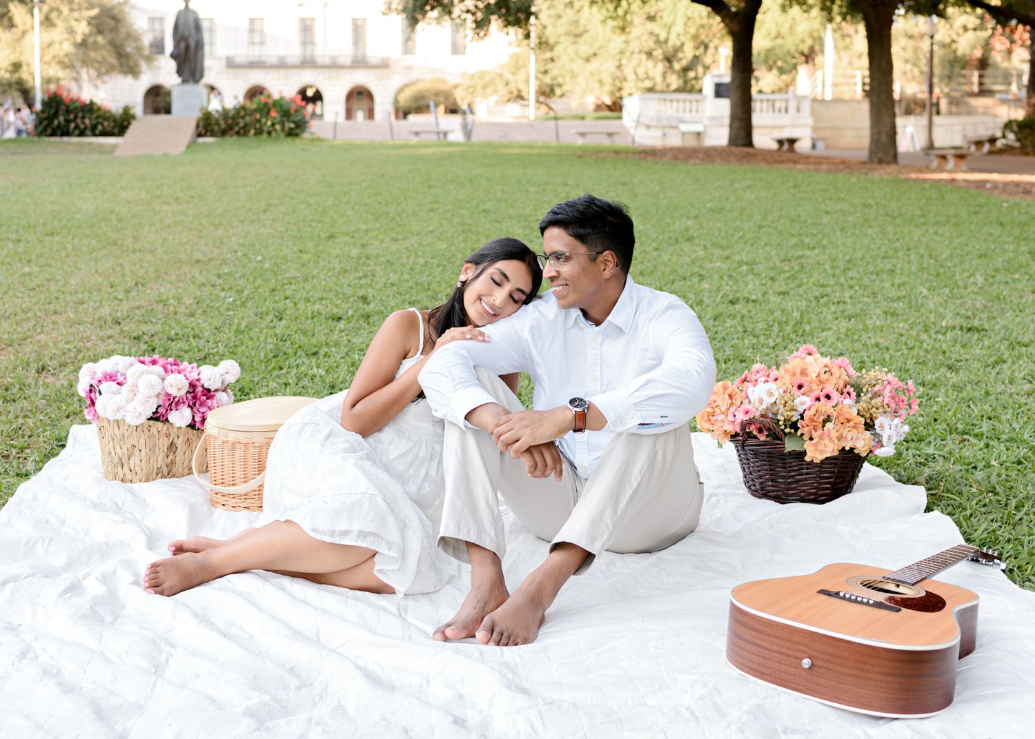 Happy couple in white outfits lounging on picnic blanket enjoying romantic outdoor date with flowers and food.